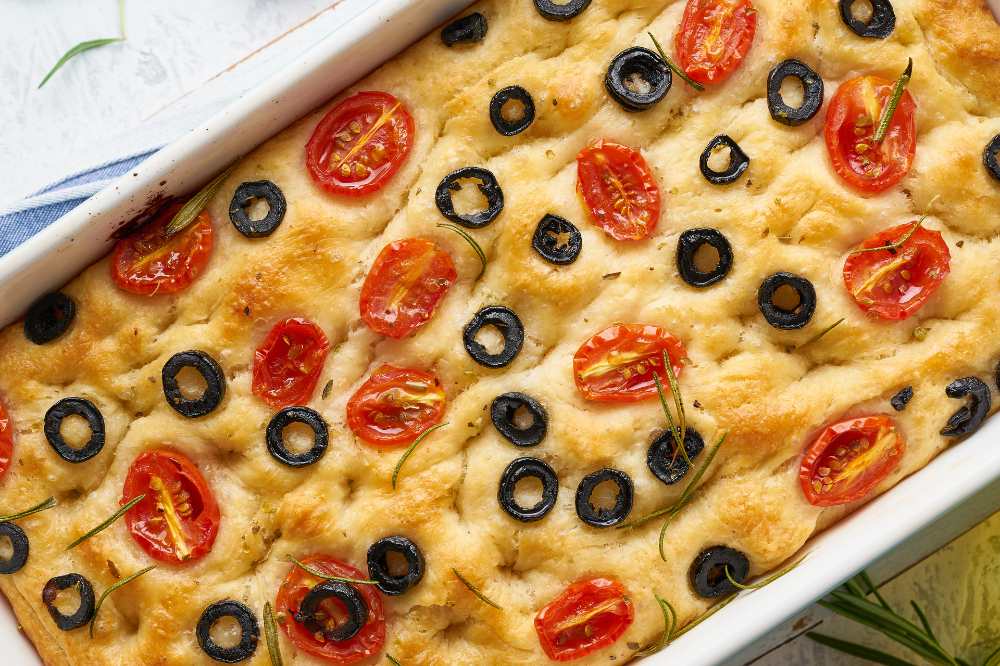 Focaccia top with half cherry tomatoes, black olives and rosemary in casserole, top view, Italian flat bread