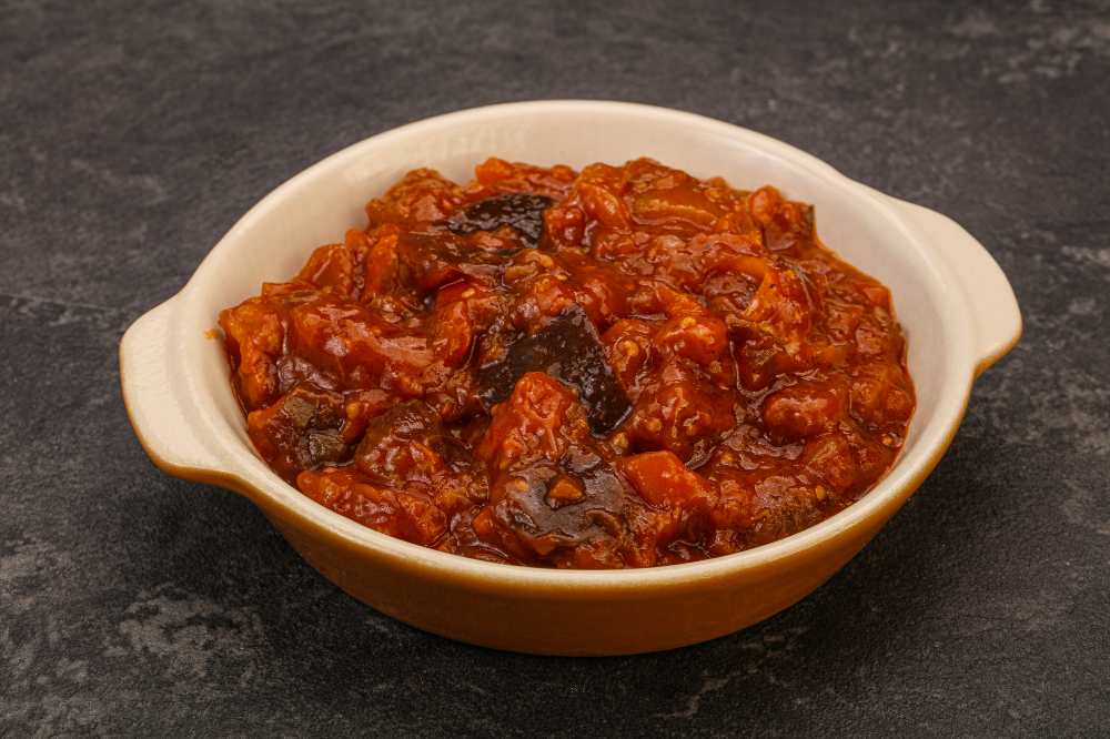 Eggplant sauté with tomato and herbs, serve in a round baking dish, on a black rustic background.
