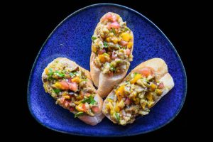 Vegetarian Bruschetta with tomatoes, eggplant, bell yellow pepper and olive oil on blue plate close up, on a black background