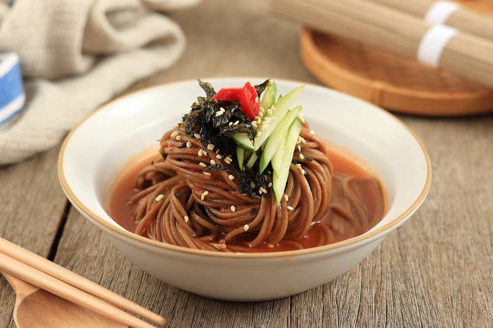 Bibim Guksu Soba Noodle, Buckwheat Cold Noodle Soup, serve in a white bowl with wooden spoon on a wooden table, top close-up view