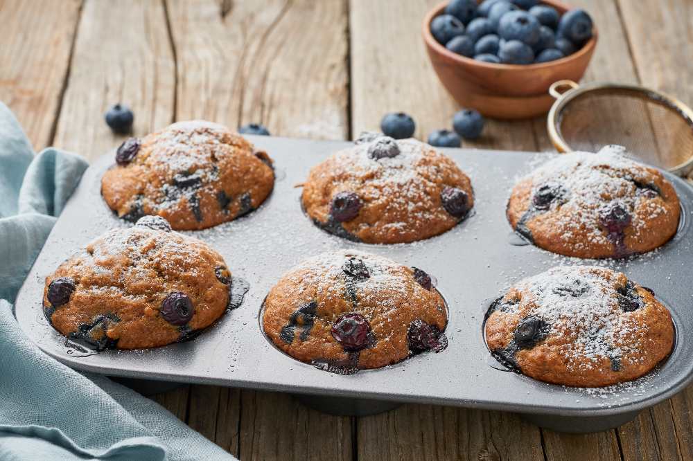 Blueberry Muffins, in a bake muffin tray with fresh blueberry on the side top with powder sugar , serve on a wooden table , close=up side view.