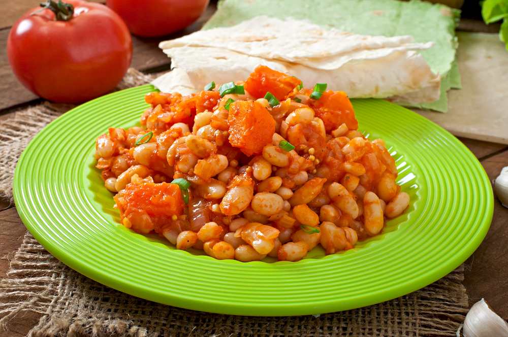 Stewed white beans and sliced pumpkin in tomato sauce, serve in a green plate with fresh tomato on the site.