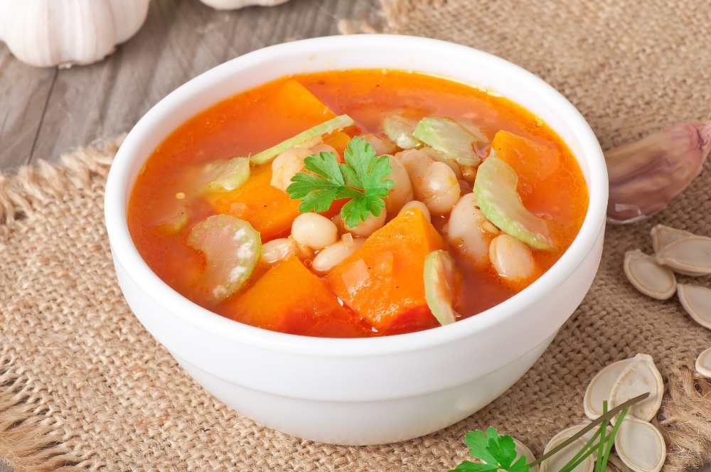 Tomato soup with pumpkin, beans and celery. serve in a white bowl with pumpkin seeds on the side and fresh garlic.