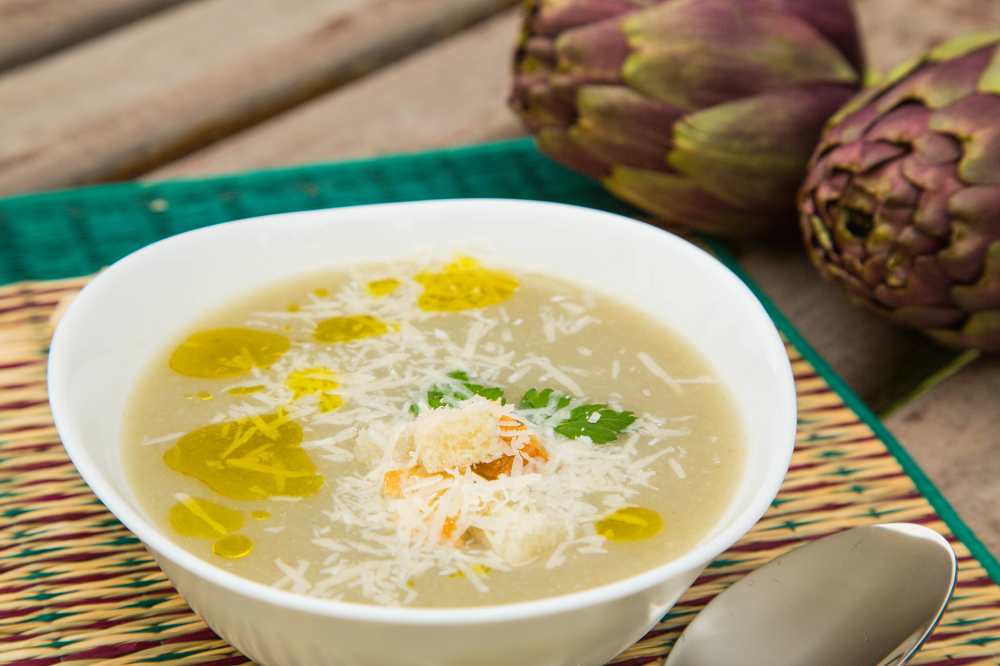 artichoke cream soup serve in a white bowl on bamboo mat with side of artichoke