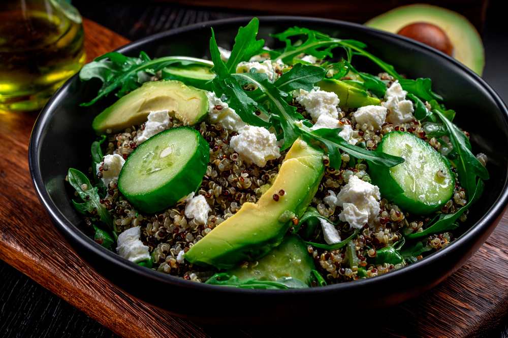 quinoa, sliced avocado, cucumber, Arugula and feta cheese. Served in a rustic black bowl, with Olive oil and half avocado on the side, serve on a wooden table.