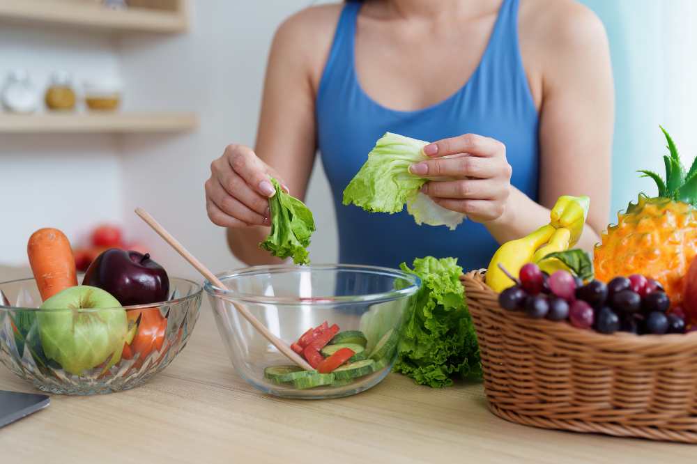 A Women in a workout cloth making salad , with fruit and vegetable on the side in a kitchen setting
