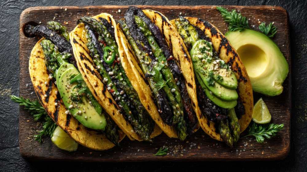 Grilled Asparagus and Avocado Tacos serve on a cutting board sit on a rustic black counter, with half avocado on the side, close-up top view.