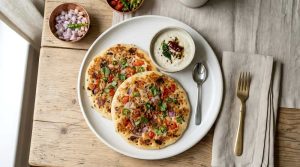 Indian Uttapams Topped with Onions, Tomatoes, Chilies, Served with Fresh Coconut Chutney and diced red onion, serve in a white plate with a small spoon and fork on the side, top view.