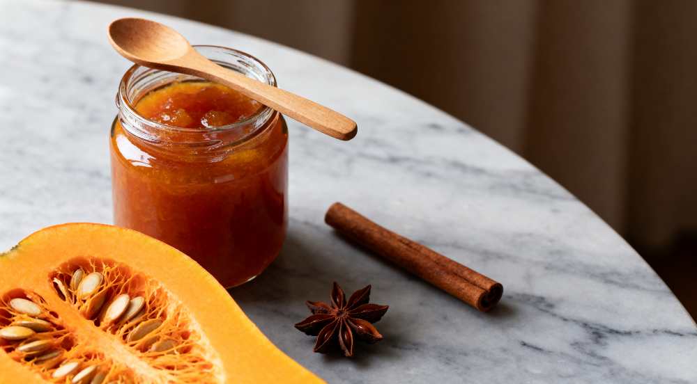 Pumpkin jam in glass jar with wood spoon on top, aromatic anis, cinnamon and raw pumpkin, close-up top view.
