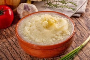 Creamy cauliflower puree serve in a brown bowl, with fresh tomato, garlic and green onion on the side, close up top view.