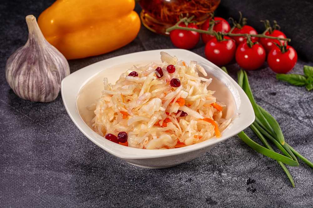 Sauerkraut salad with cranberries and carrots serve in a white bowl, with fresh garlic, yellow pepper, cherry tomato and green onion on the side, close-up top view.