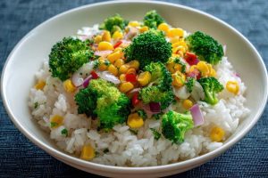 Steamed Rice Topped with Broccoli Florets Corn Kernels and Red Onion Chunks, serve in a white bowl in a blue background , close up top view.